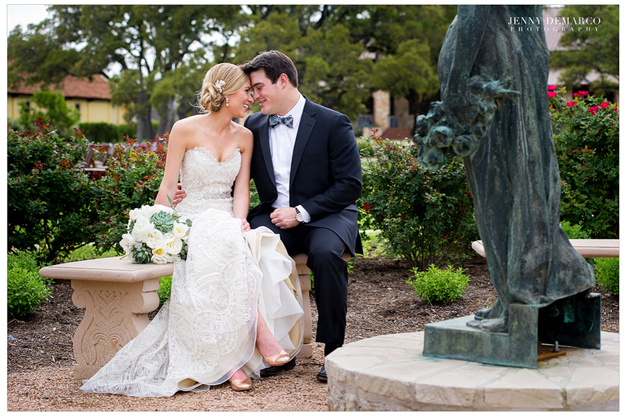 The bride and groom are sitting on a bench together in a garden with a copper statue.