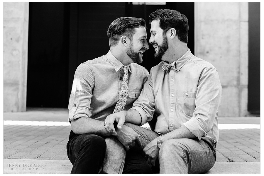 Black and white photo of cute gay couple sitting on the sidewalk and laughing.