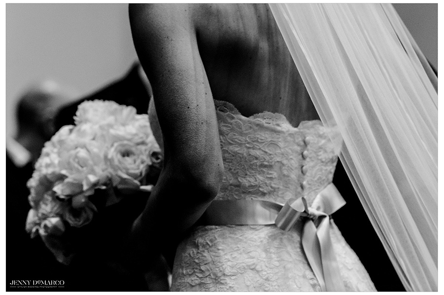 A black and white image centered on the aesthetically pleasing details of the bride's dress, flowers, and veil. The lace and buttons on the bodice of the dress contract with the silk of the bow on the back of the dress. 