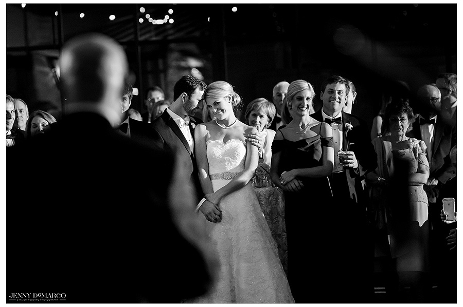 The groom embraces the bride as she looks loving at her father who is giving them a toast. This black and white image centers on the appreciation the bride has for her father.