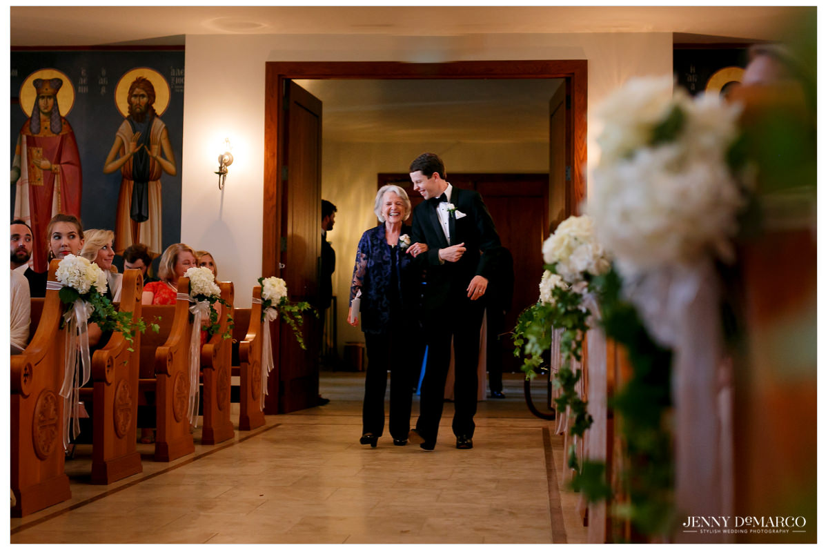 Groom's mom walks her son down the aisle while laughing. 