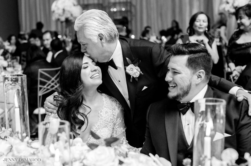 Father of the bride kissing his daughter on the head and hugging his new son in law in black and white candid moment.