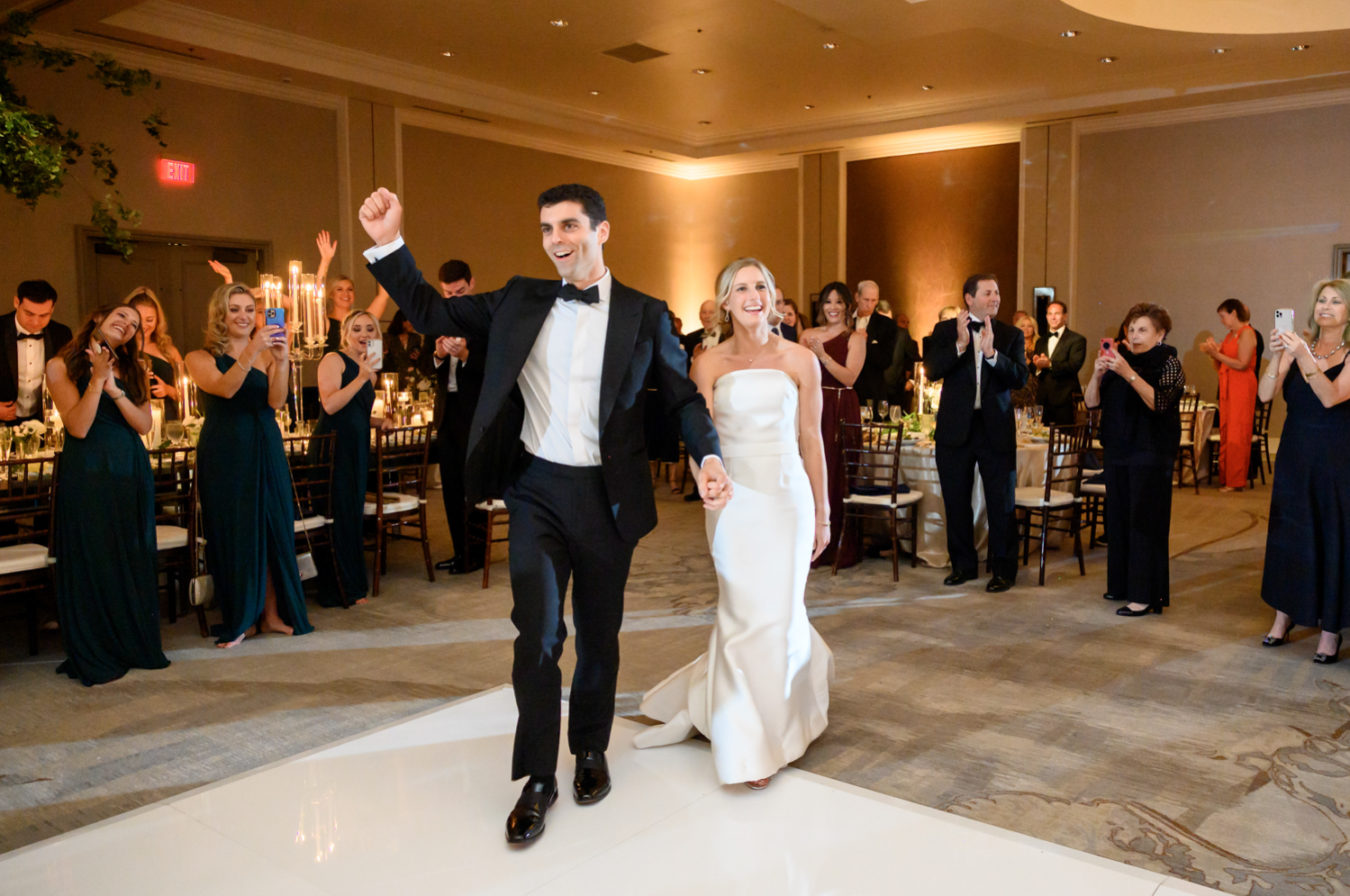 The bride and groom enter the reception as the guests applaud. The groom pumps his fist in the air.