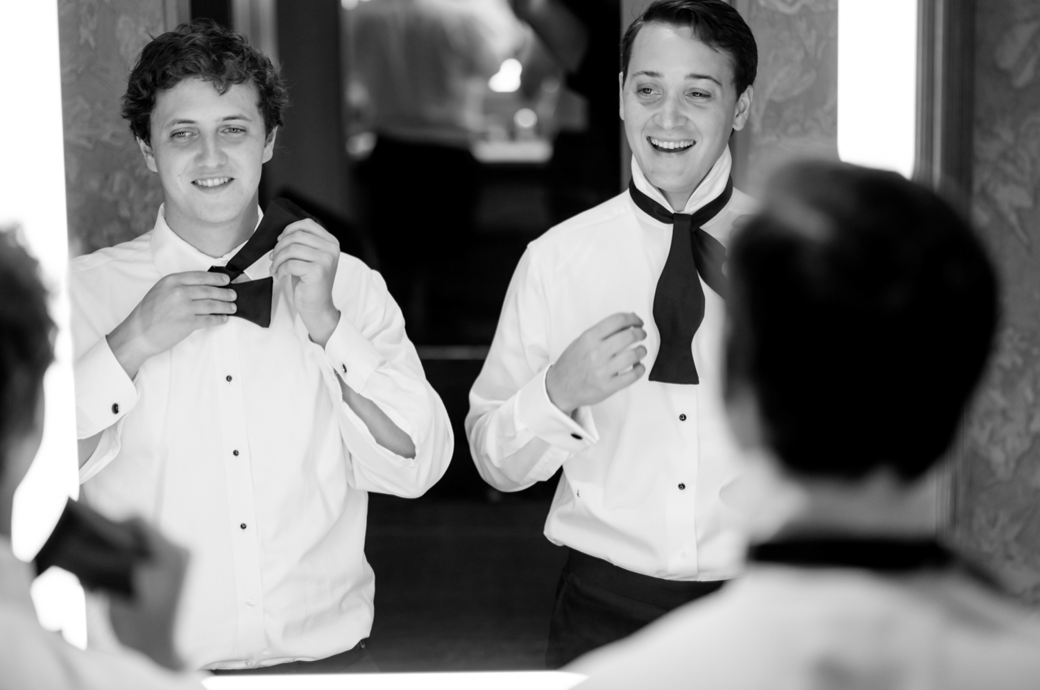 The groom and his brother tie their bowties in the mirror and laugh together.