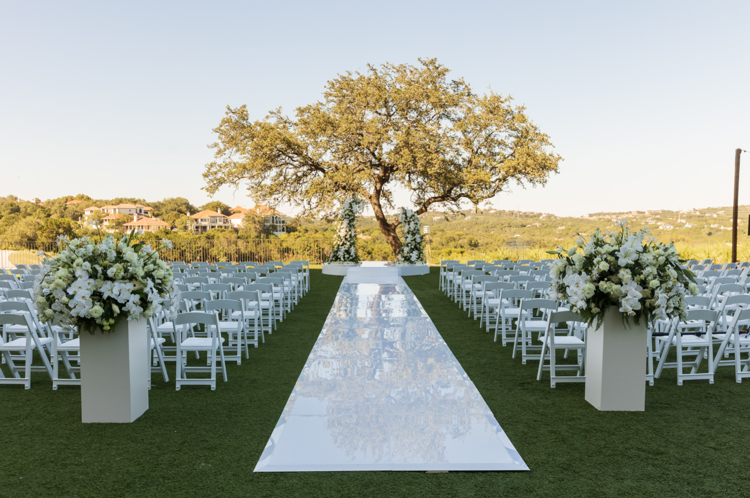 A shiny, acrylic aisle sits between rows of chairs and leads to an alter covered in white flowers.