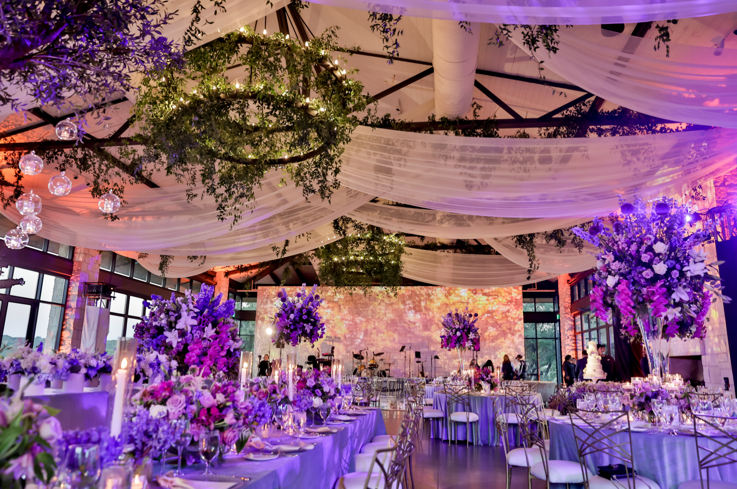 A shot of the reception details in the blue mood lighting. Greenery hanging from the chandeliers, sheets of tulle draped across the ceiling, large flower arrangements, sparkling dinnerware, and blue table linens decorate the room.