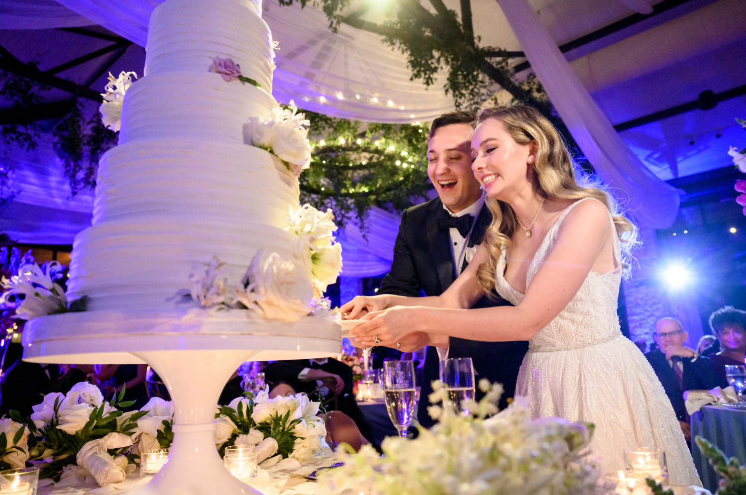 The bride and groom cut into the 5-tier wedding cake, decorated with white flowers.