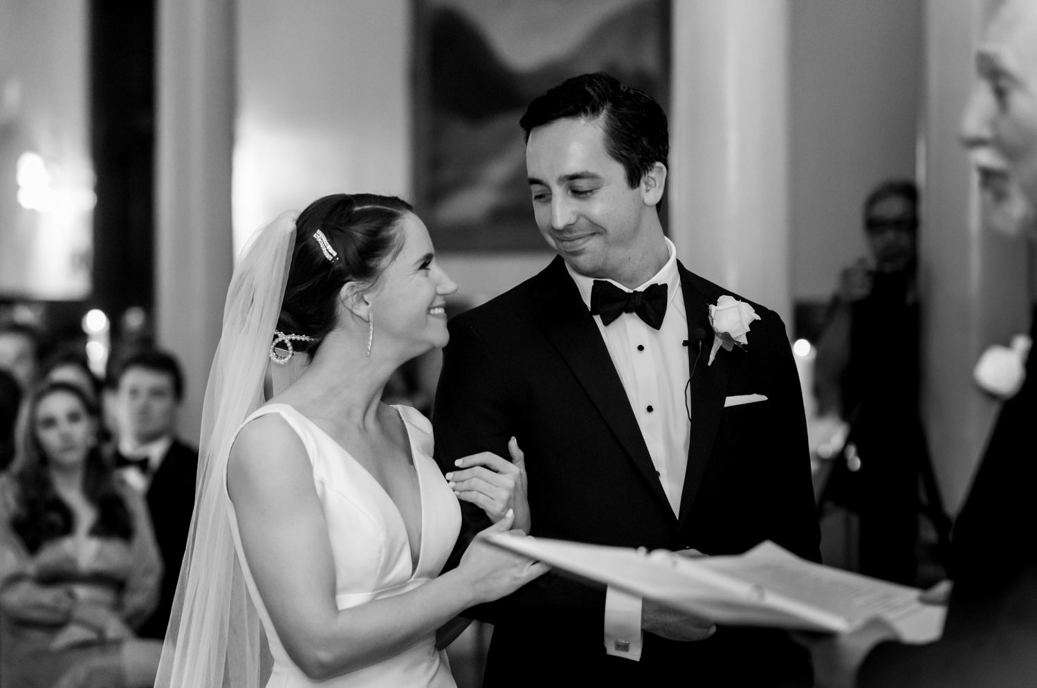 The bride holds the grooms arm and they smile at each other, standing at the altar