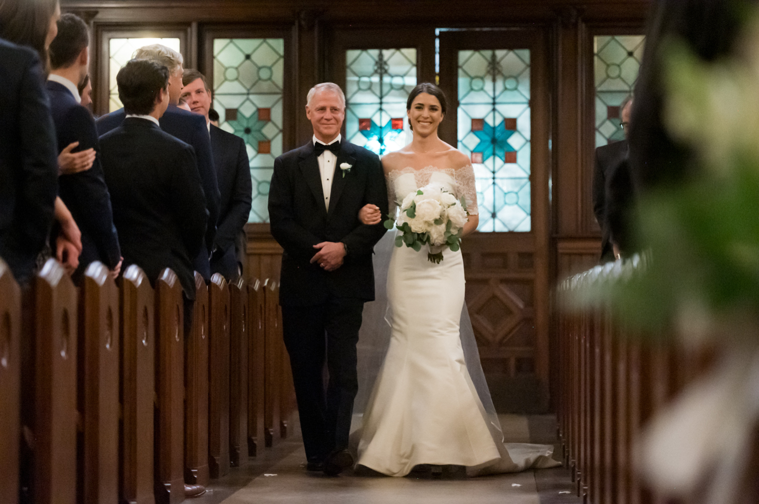 The bride walks down the aisle with her father, smiling at their guests.