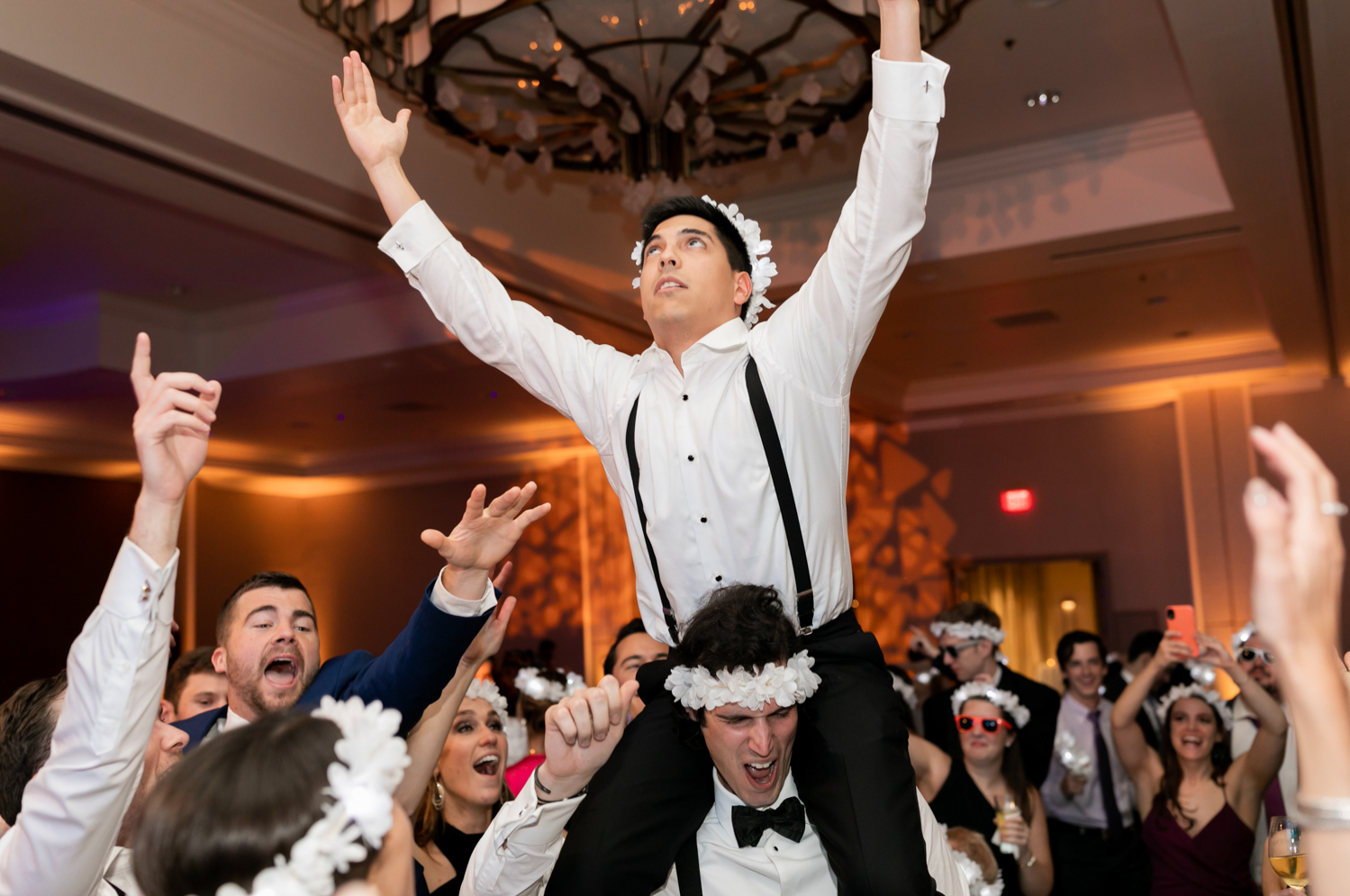 A guest sits on his friends shoulders, wearing flower crowns and dancing with friends at the reception.