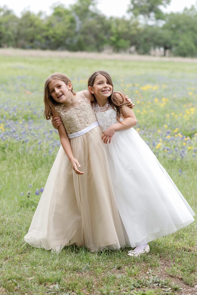 flower girls in their white and champagne dresses smiling and hugging in a field of wildflowers