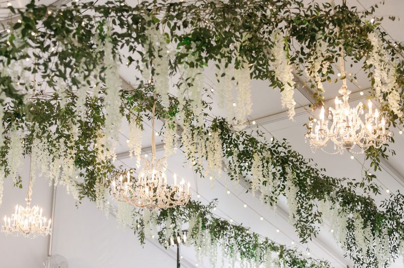greenery and white florals hanging from the ceiling of the tent with gorgeous chandeliers