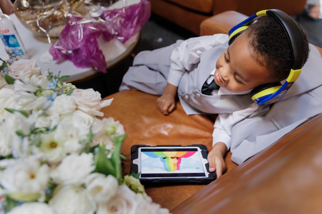A lighthearted image of a young flower girl momentarily distracted by an iPad, with a bouquet of white and blush roses resting beside her.