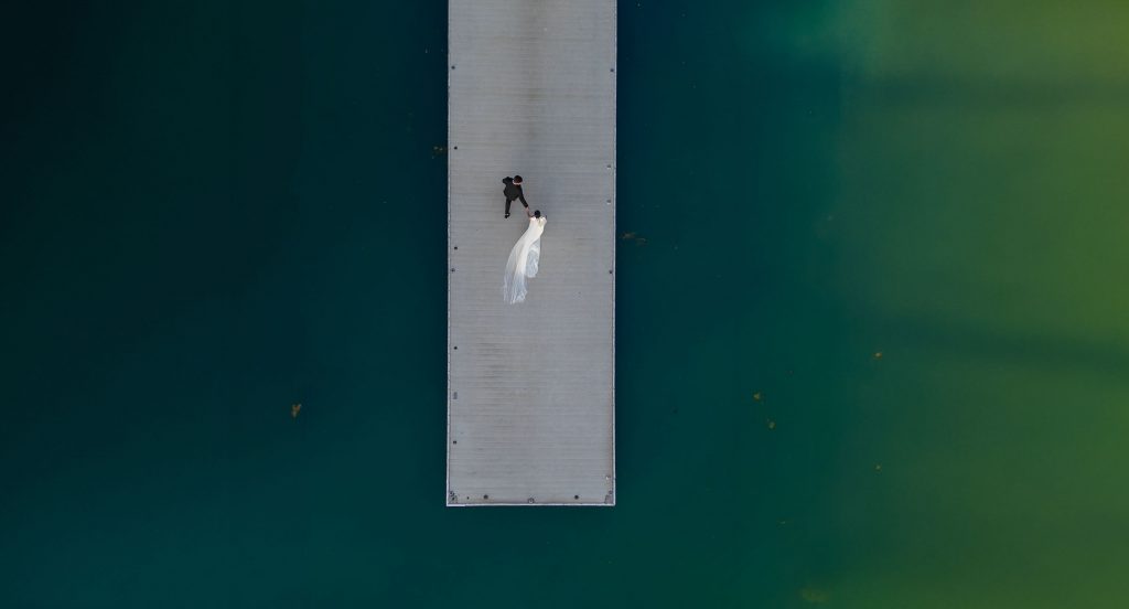 Aerial portrait of a wedding couple standing on a dock surrounded by blue water