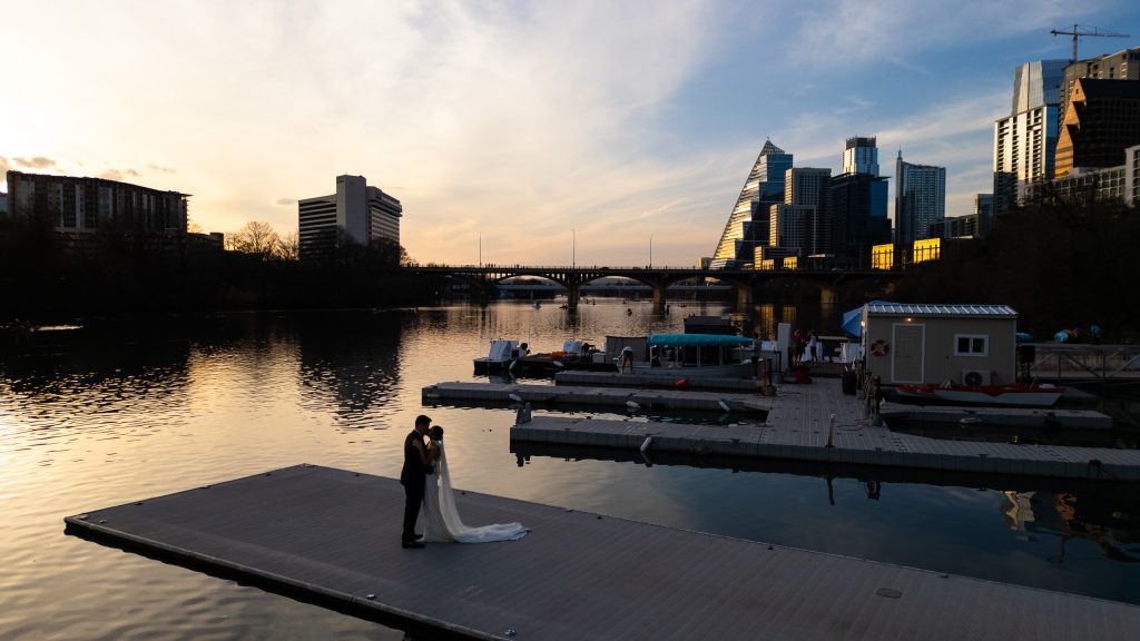 Bride and groom standing beside the water with the Austin skyline at sunset