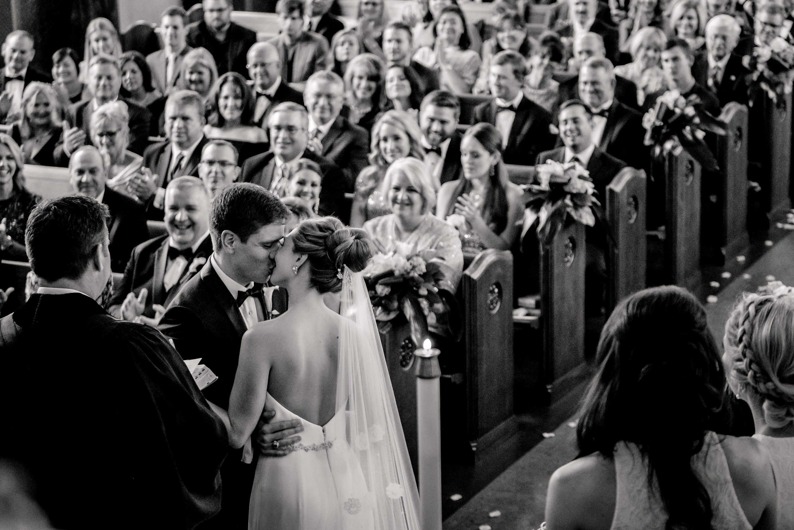 Black and white photo of a newly married couple sharing a kiss as guests celebrate around them