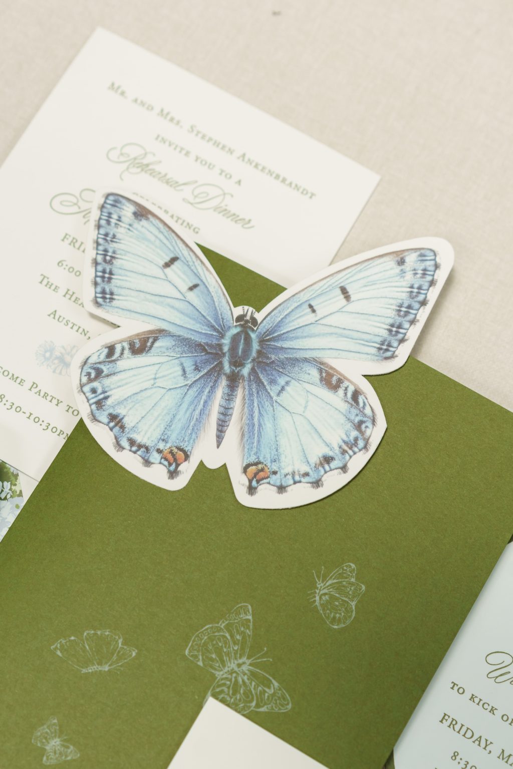 Illustrated blue butterfly placed over a traditional green and ivory wedding invitation suite