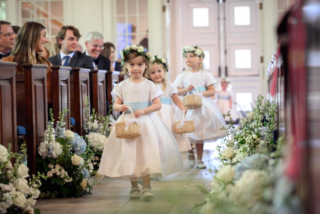 Young flower girls walk through the blue and white floral aisle during the church ceremony wearing floral crowns