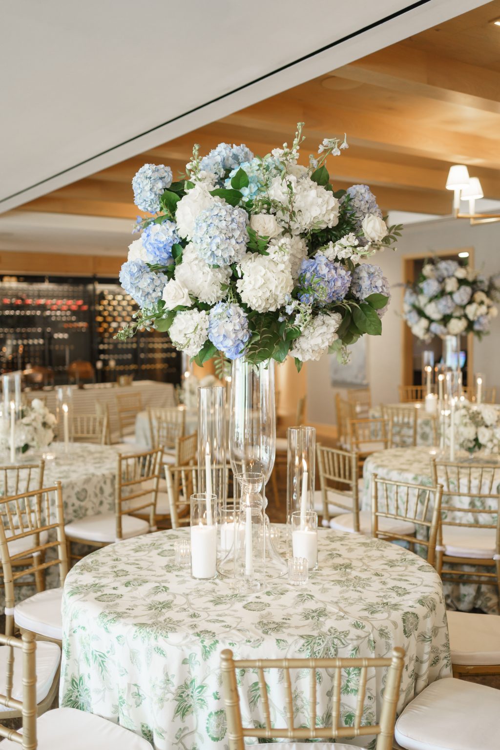 Tall blue and white hydrangea floral centerpiece surrounded by patterned linens and gold chairs