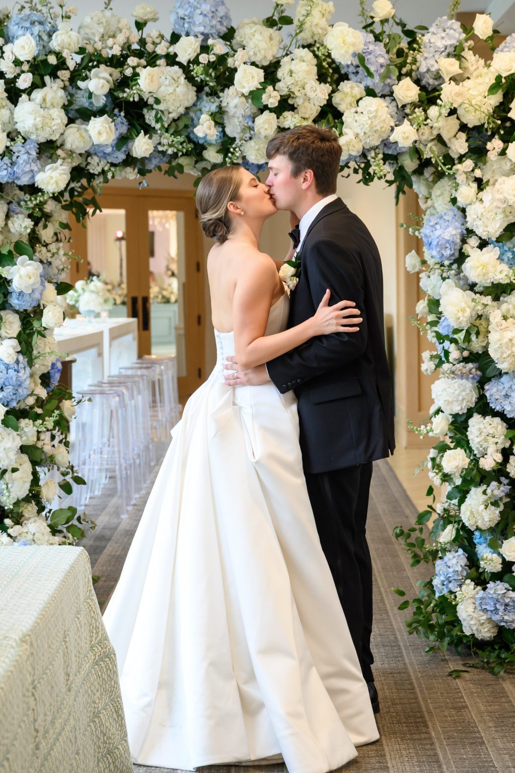 Newlyweds kiss beneath an abundant blue and white floral arch at Westwood Country Club