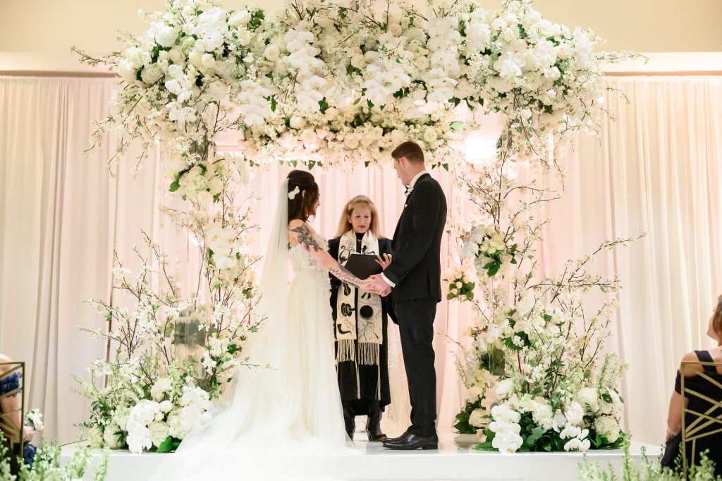 Bride and groom standing beneath a white floral chuppah inside the Four Seasons Austin Ballroom